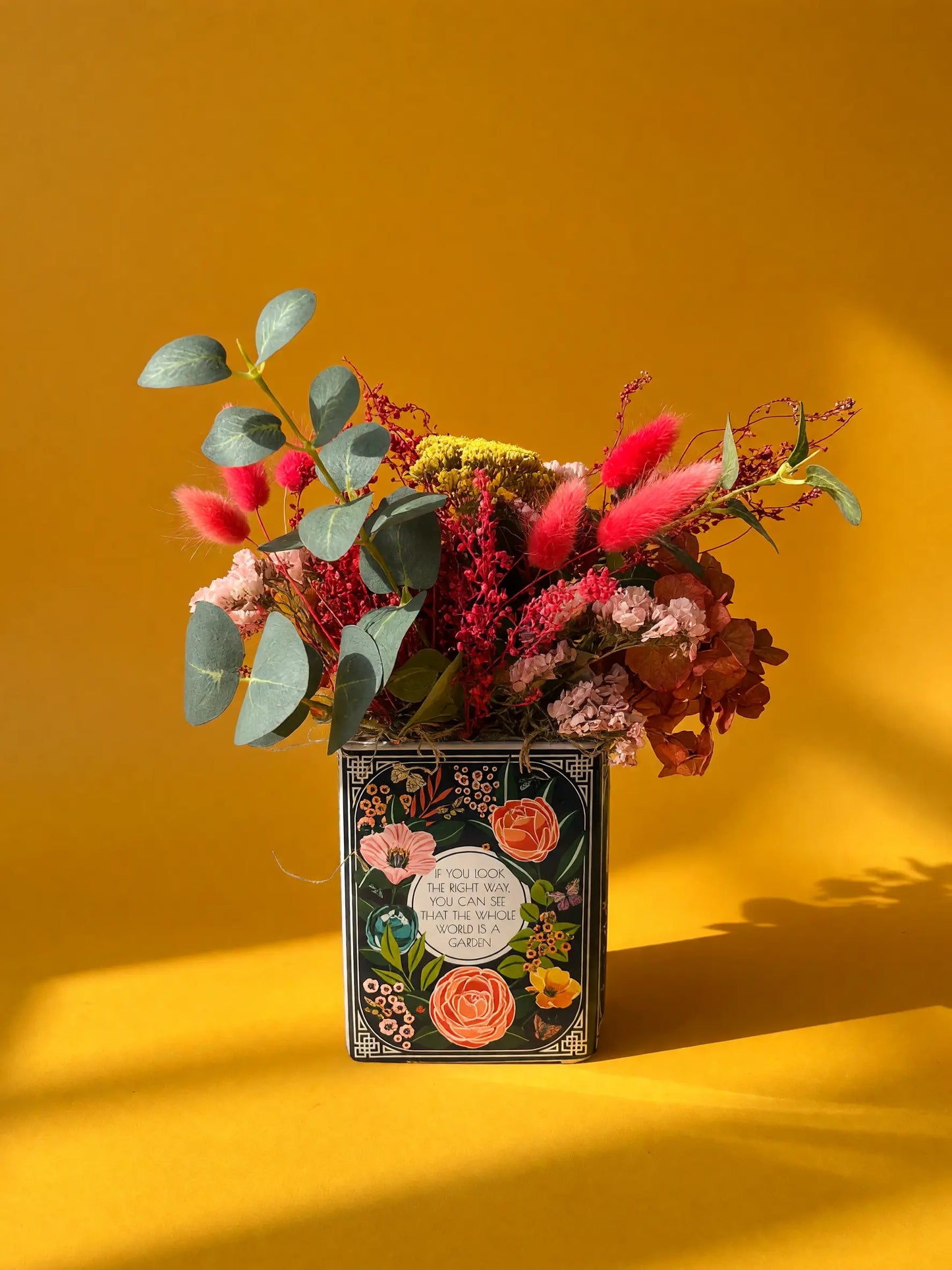 A decorative vase filled with a colorful dried flower arrangement, set against a solid mustard yellow background with soft shadows.