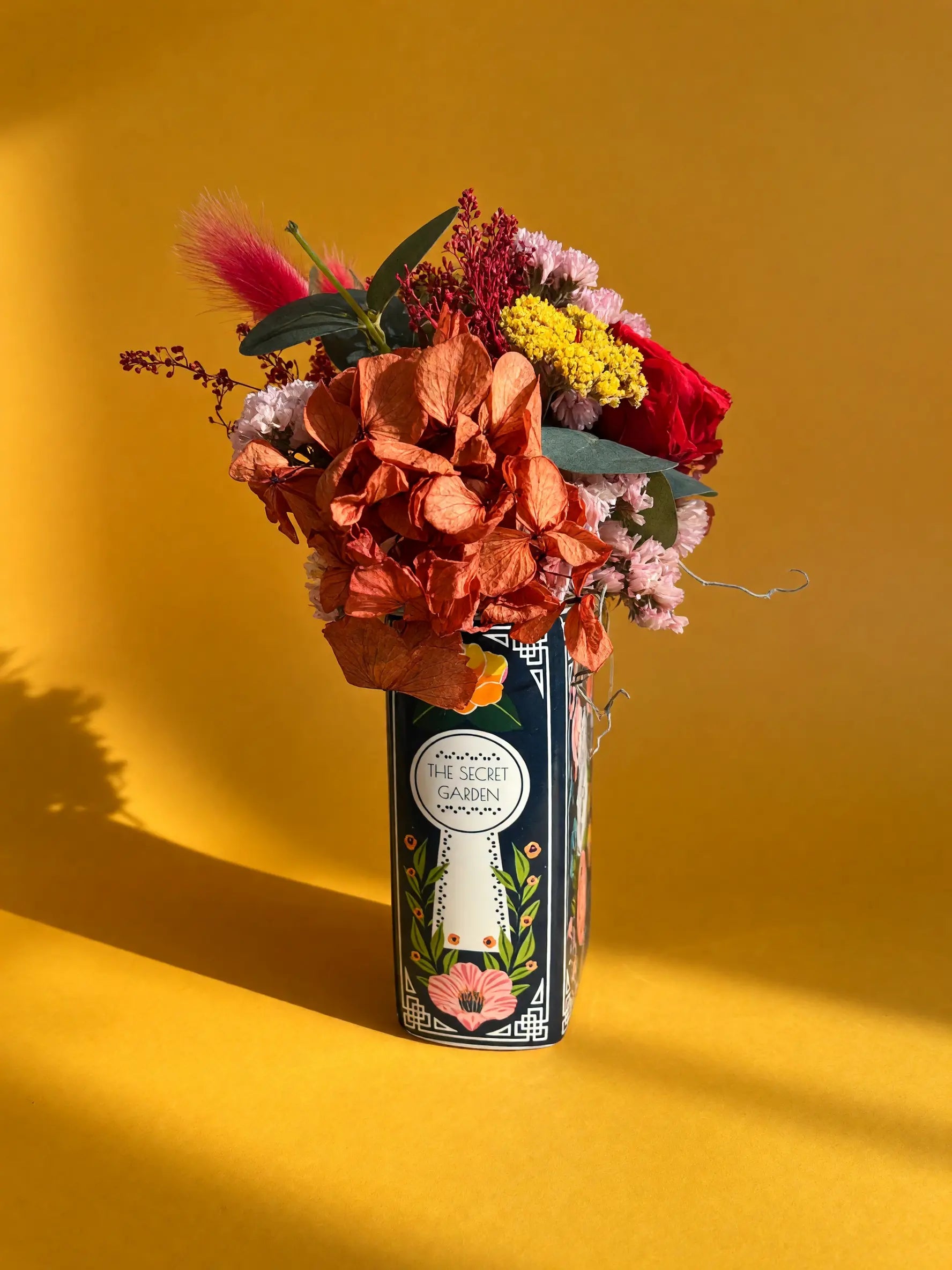 A decorative cylindrical vase with a floral label reading 'THE SECRET GARDEN' holds a colorful dried flower arrangement against a warm yellow background.