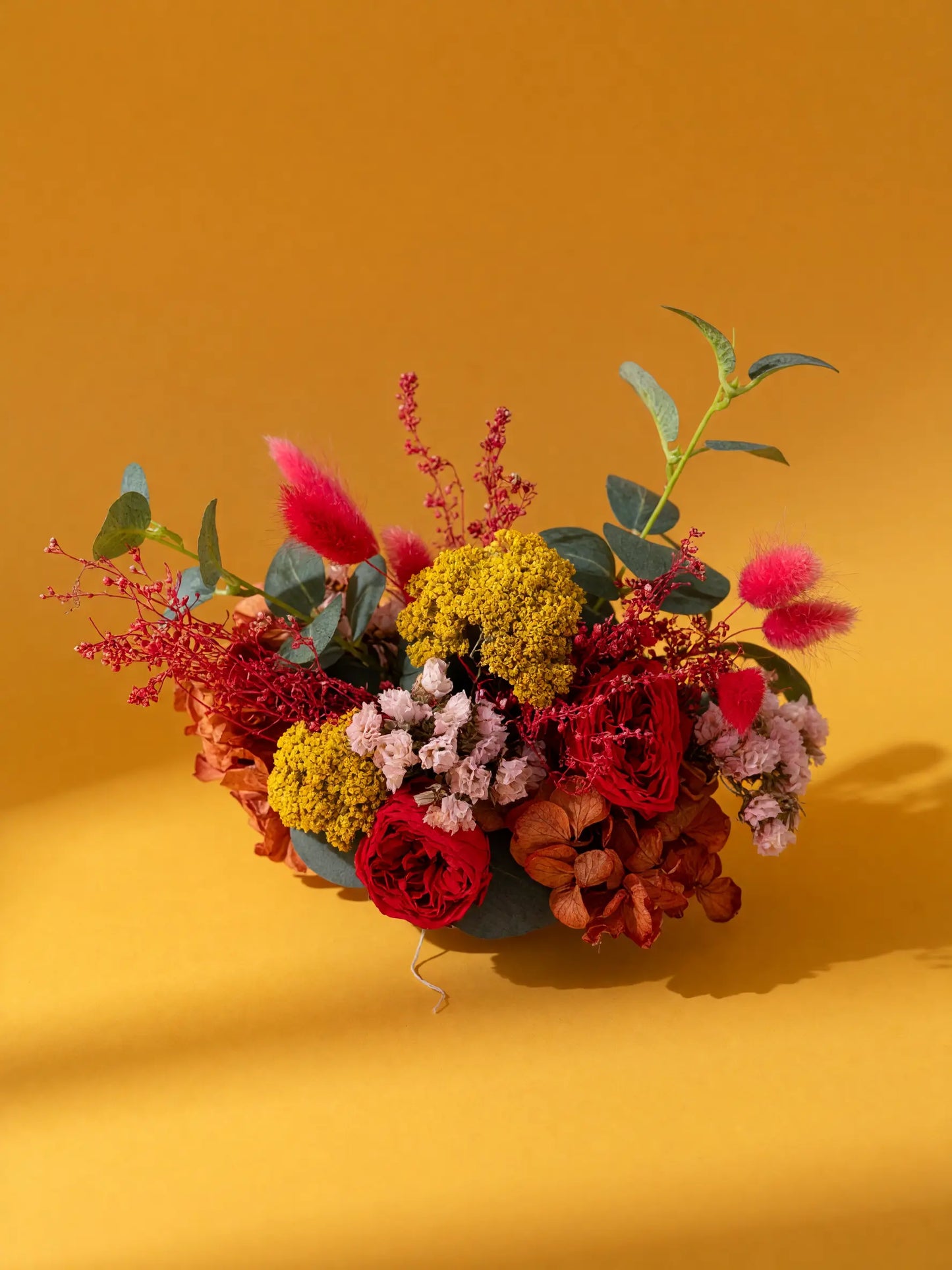 A vibrant artificial floral arrangement in a dark green bowl, featuring red roses, yellow clusters, pink fluffy stems, and green leaves, set against a solid mustard yellow background.