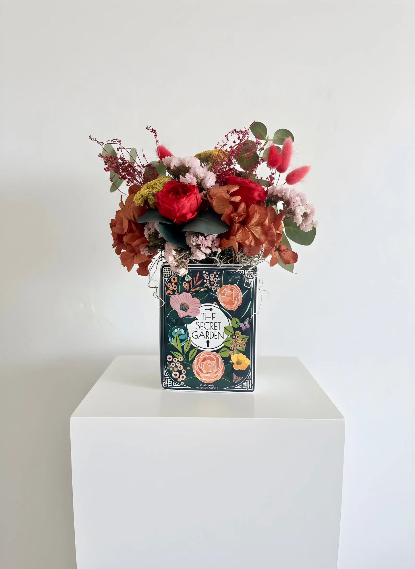 A decorative tin vase labeled 'The Secret Garden' filled with a colorful faux floral arrangement, displayed on a white pedestal against a white wall.